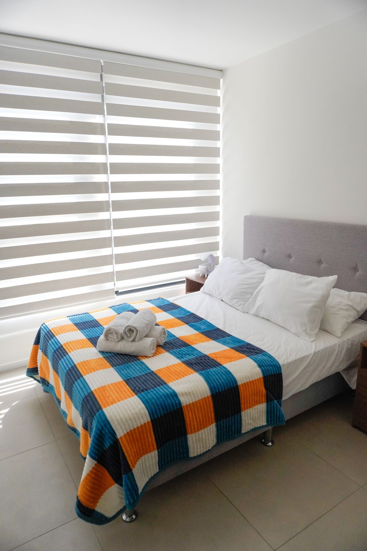 Modern bedroom with striped blinds, colorful checkered bedspread, and rolled towels on the bed.