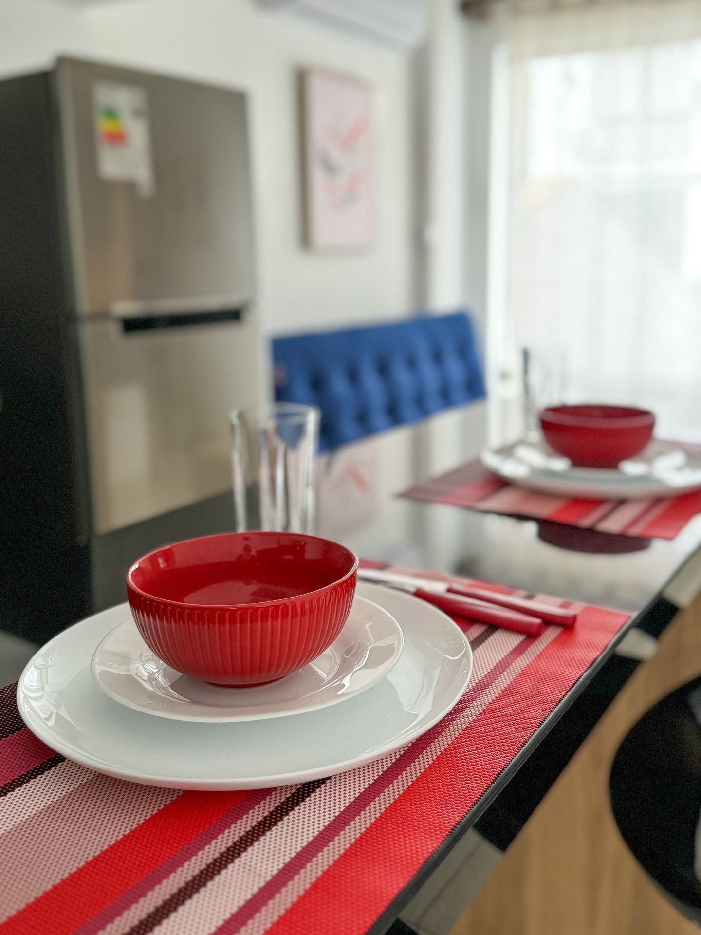 Table setting with red bowl on white plates and red placemat in front of a refrigerator.